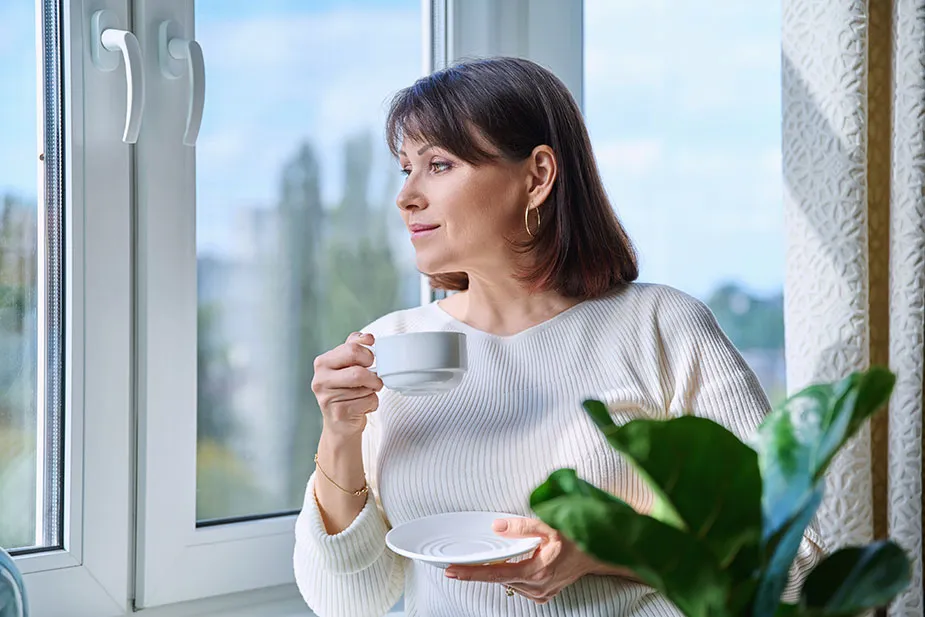 Žena srednjih godina pije čaj i gleda kroz prozor – razmišljanje o ranoj menopauzi.