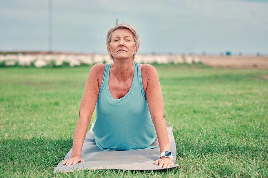 Zrela žena izvodi pozu joge na prostirci u prirodi, simbol fizičke aktivnosti i mentalnog balansa u menopauzi.