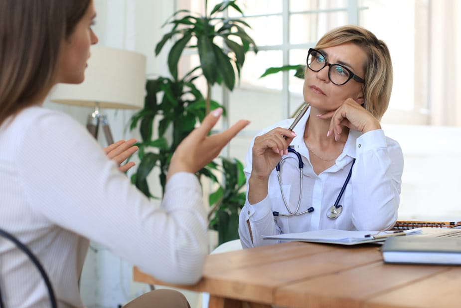 Žena srednjih godina razgovara sa doktorom u savremeno opremljenoj ordinaciji, dok doktor pažljivo sluša i razmišlja o njenim rečima.